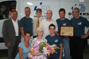 Group of Demers employees and family members posing together indoors with an award and flowers