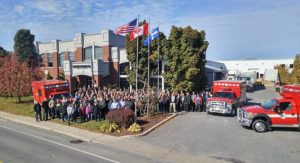Large group of people standing outside an industrial building with Demers emergency vehicles parked nearby